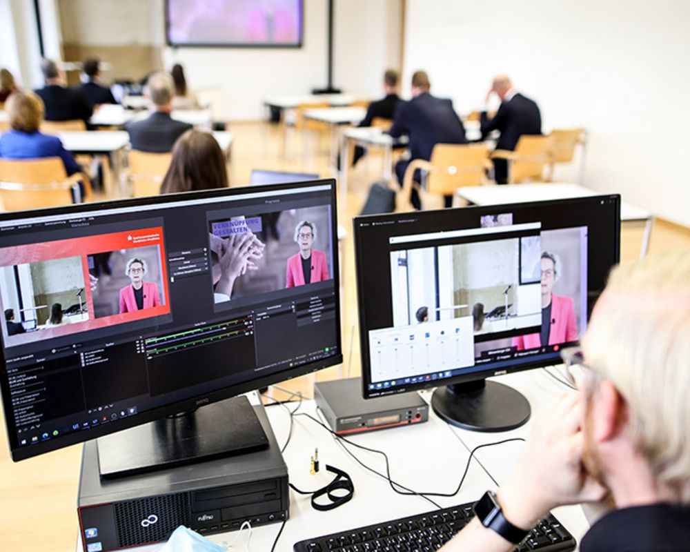 In einem Seminarraum sind im Hintergrund Teilnehmende einer Weiterbildung zu sehen, während im Vordergrund über Monitore deutlich wird, dass sich externe Teilnehmende digital zur Veranstaltung zugeschaltet haben.