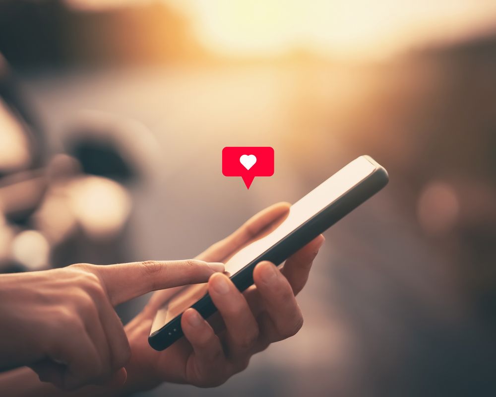 Handy wird in der Hand gehalten. Darüber erscheint ein Herz-Symbol, welches ein Like symbolisiert. Im Hintergrund ist ein Sonnenaufgang zu sehen.