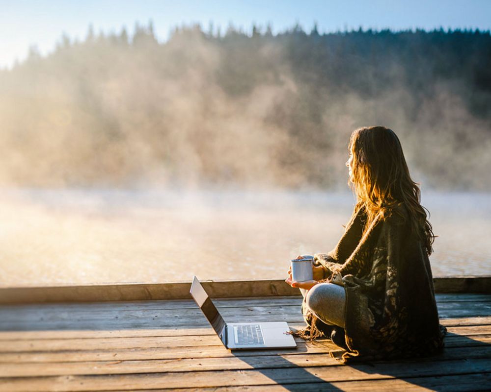 Eine Frau sitzt alleine mit ihrem Laptop auf einer Seeterrasse und lernt für eine Weiterbildung.