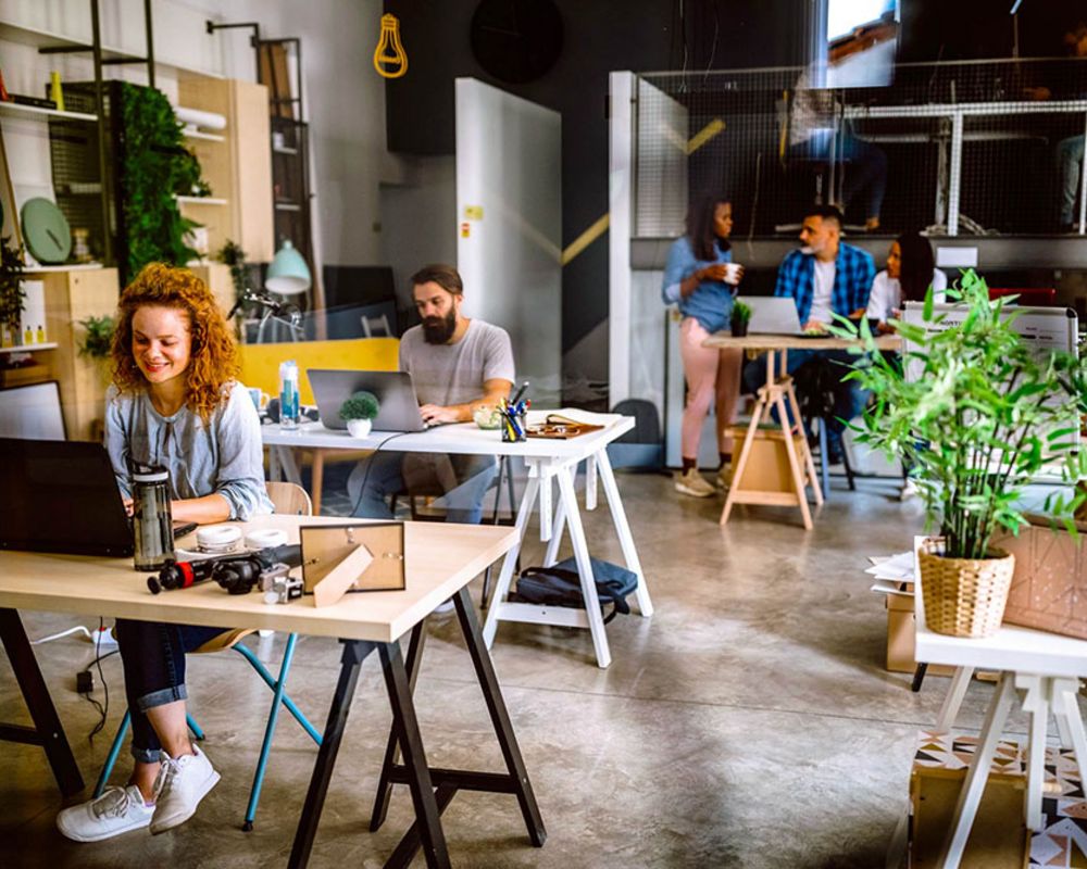 In einem modern eingerichteten Büro, welches Prinzipien der "New Work" aufgreift, sitzen Mitarbeiter an ihren Schreibtischen und stehen an der Küchenzeile.