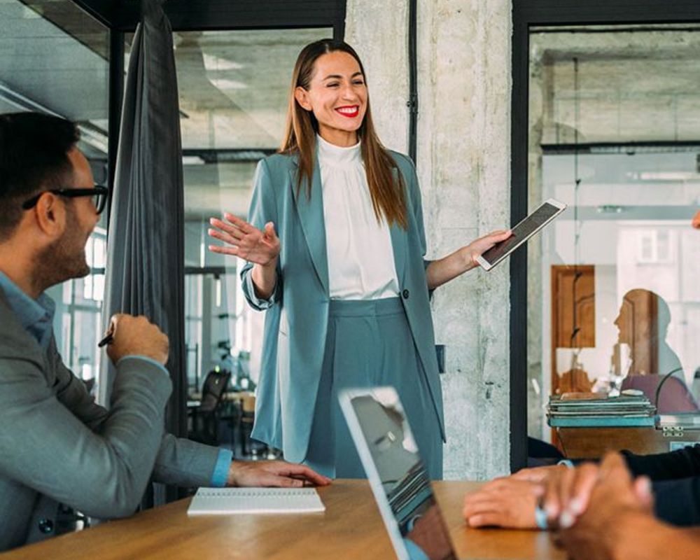 Eine Führungskraft aus dem Vorstand präsentiert eine Idee vor dem Team, welches vor ihr an einem großen Tisch sitzt und gespannt zuhört.